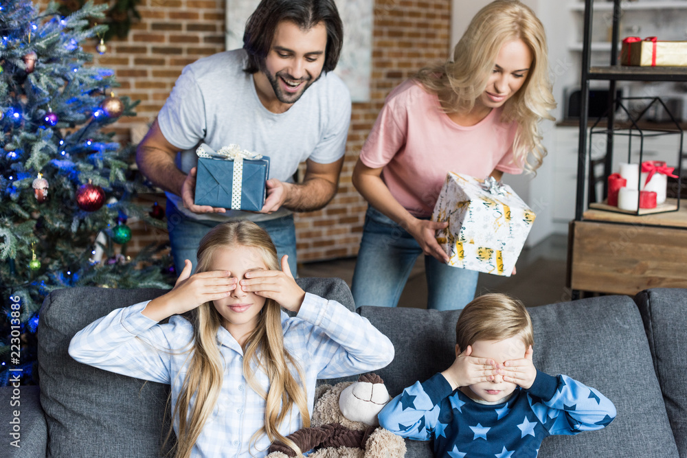 parents presenting christmas gifts to adorable children in pajamas closing eyes