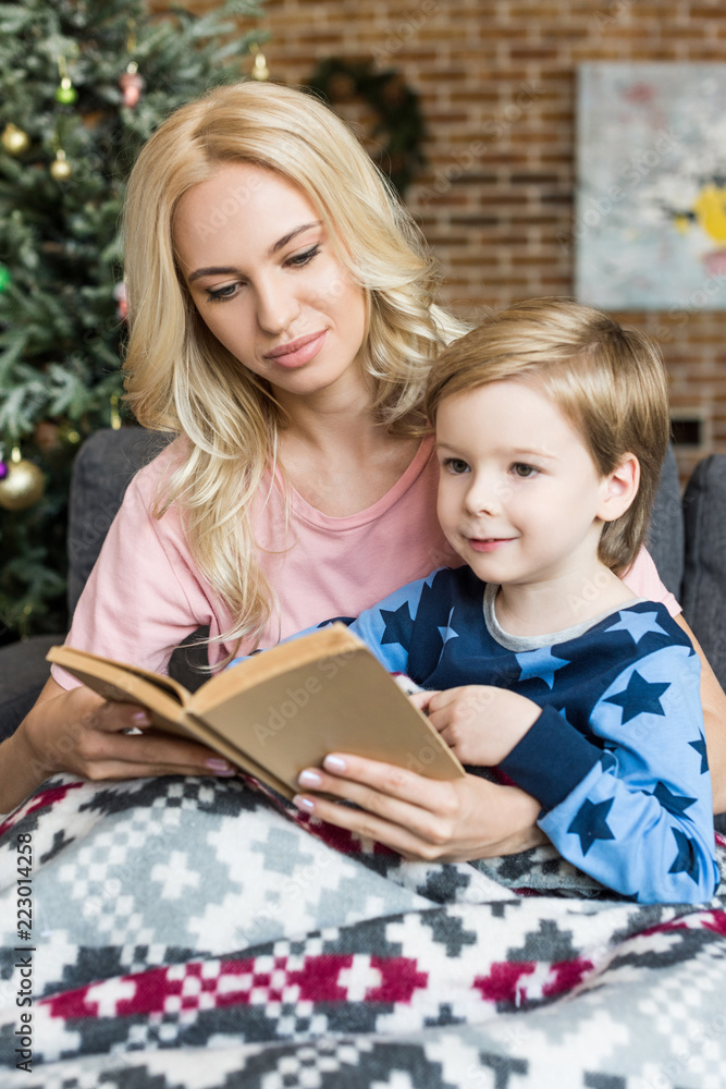 beautiful young mother and adorable smiling son reading book together at christmas time beautiful young mother and adorable smiling son reading book together at christmas time