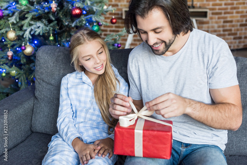 cute smiling daughter in pajamas looking at happy father opening christmas present