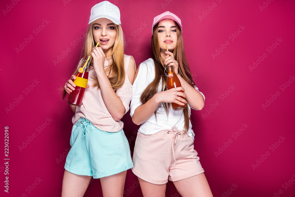 Two lovely young ladies in cute sportswear drinking cold beverages from bottles and looking at camera while standing on magenta background Two lovely young ladies in cute sportswear drinking cold beverages from bottles and looking at camera while standing on magenta background