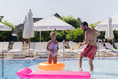 Water games. Nice handsome man standing in the pool while playing together with his daughter
