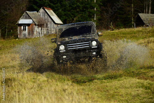 SUV crossing puddle and making splash. Off road car in black color takes part in racing. Extreme and four wheel drive concept. Cross country rallying or rally raid near countryside hut aside forest