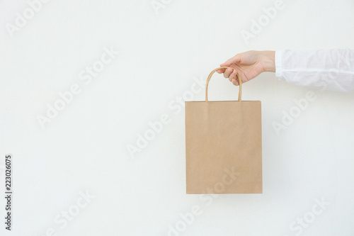 Young caucasian woman holds in hand empty blank mock up brown craft paper bag on white wall background. Christmas New Years present shopping sale. Conceptual. Banner poster with copy space