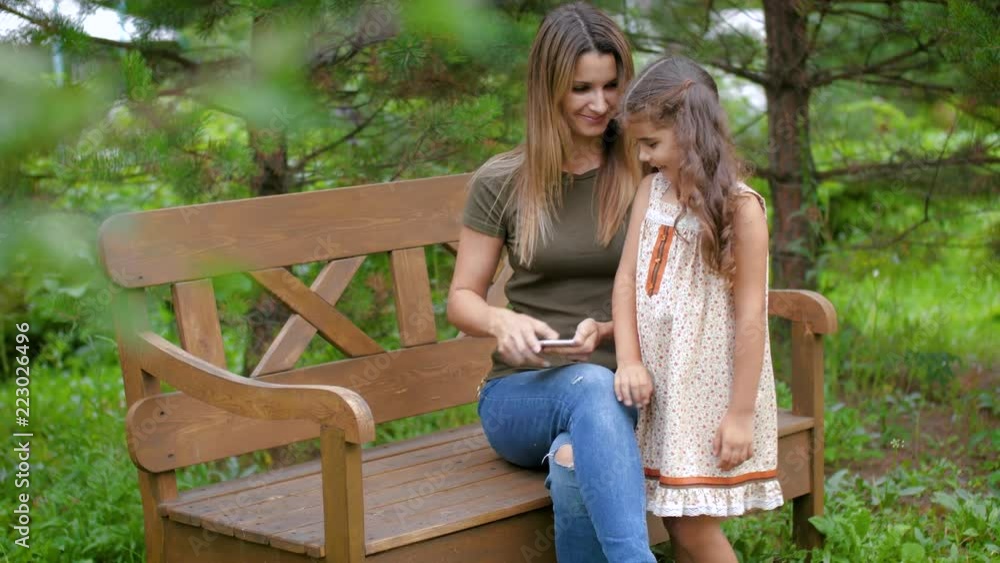 The woman sitting on a bench removes the smartphone after arrival of the child. The girl comes to her mother and sits down on her knees. The woman sitting on a bench removes the smartphone after arrival of the child. The girl comes to her mother and sits down on her knees.