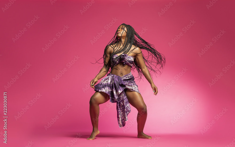 Beautiful African Black girl wearing traditional colorful African outfit does a dramatic dance move against a colorful pink background