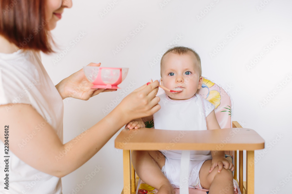Mother Feeding Baby Sitting In High Chair At Mealtime Mother Feeding Baby Sitting In High Chair At Mealtime