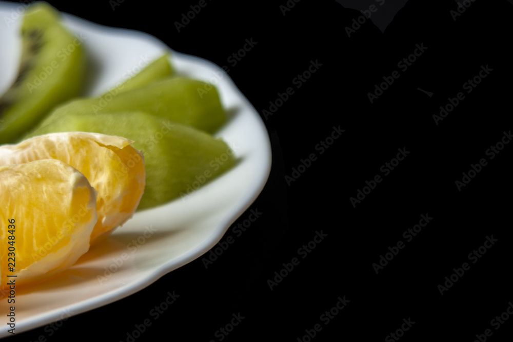 Kiwi and mandarin orange slices placed on white plate in half, aligned at left, isolated on black background. Space for text.