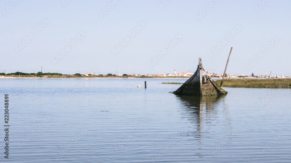 Single and lonely sunken boat half-submerged in the estuary of Aveiro Lagoon, Portugal. Small quaint town in horizon.