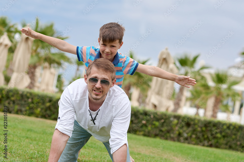 Obraz premium Cute european boy is sitting on his fathers back at the green grass of the resort. His arms are open wide and he is looking to the camera.