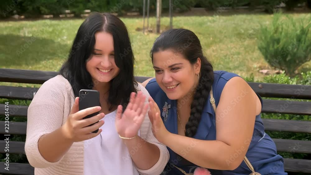 Two young obese girls waving in camera on smartphone and smiling with relax on the bench in park Two young obese girls waving in camera on smartphone and smiling with relax on the bench in park