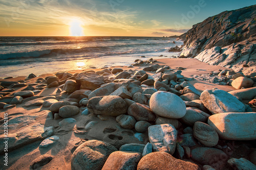 une plage avec des pierres en premier plan face à un coucher de soleil sur l'océan et des falaises