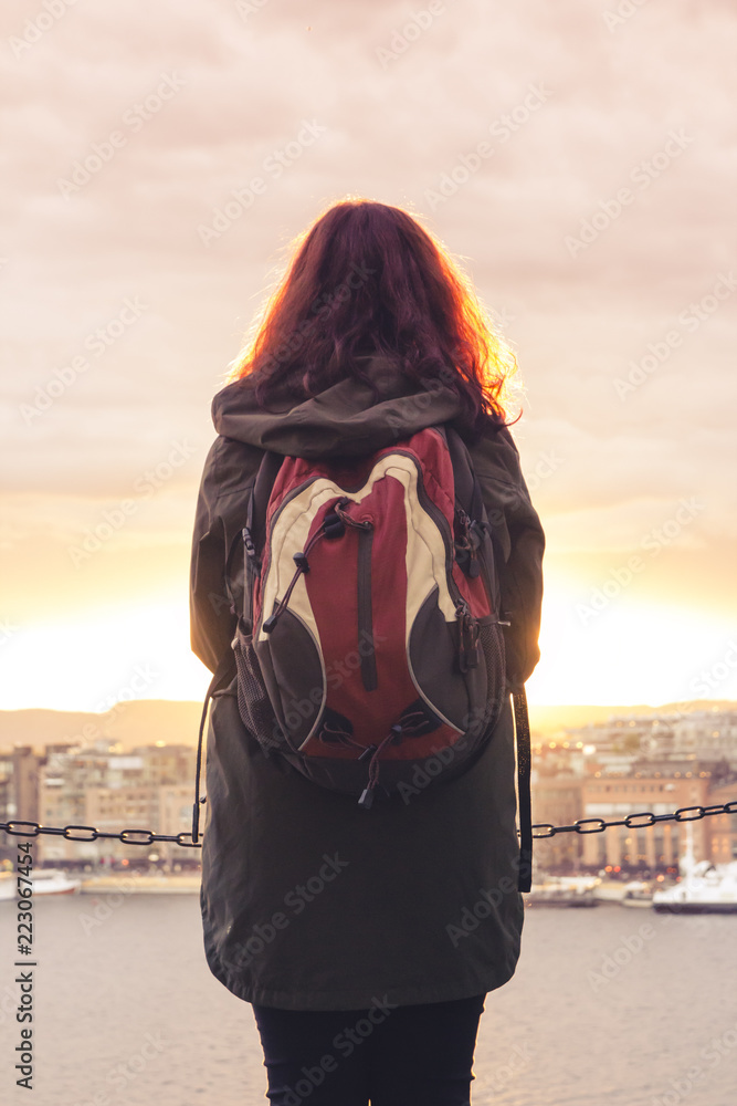 A lonely girl with a backpack stands with her back and looks at the beautiful panorama of the city and the river at sunset, concept of tranquility, travel and serenity