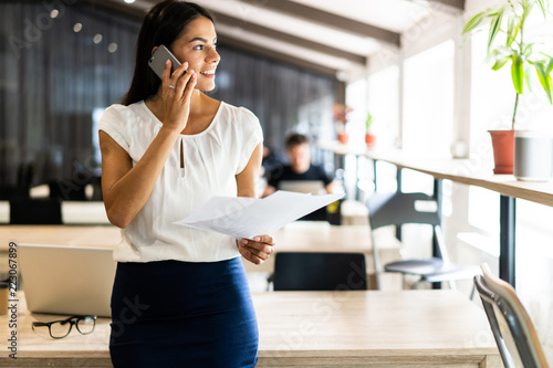 Everyday paperwork. Pensive young beautiful businesswoman in glasses talking on mobile phone and looking through documents while standing near window.