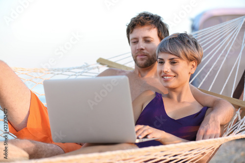 Young couple resting with laptop in hammock on beach