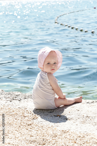 Adorable baby girl on the beach, wearing a white bodysuit and pink striped bonnet, summer vacation concept