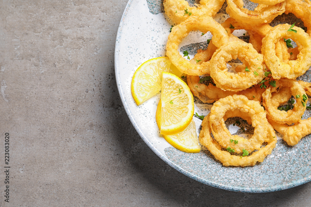 Plate with homemade crunchy fried onion rings and lemon slices on gray background, top view. Space for text