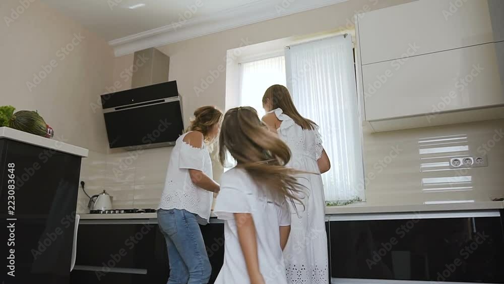 Happy little girl in white dress with her sister and mom are dancing and listen music while cooking eating in kitchen at home. The cute girl dances around herself her mom with sister prepare food in Happy little girl in white dress with her sister and mom are dancing and listen music while cooking eating in kitchen at home. The cute girl dances around herself her mom with sister prepare food in