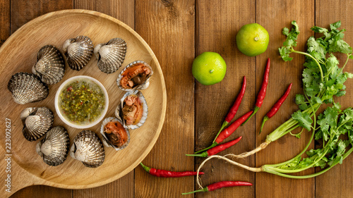 Flat lay of boiled cockles in wooden plate with chilli dipping sauce on wooden table with red chilli pepper lemon and pak chee