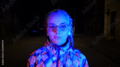 Attractive pretty girl with unusual hairstyle walking on street with light at night. Dyed blue hair in braids. Pensive hipster teenager in glasses chews cud and blows bubbles.