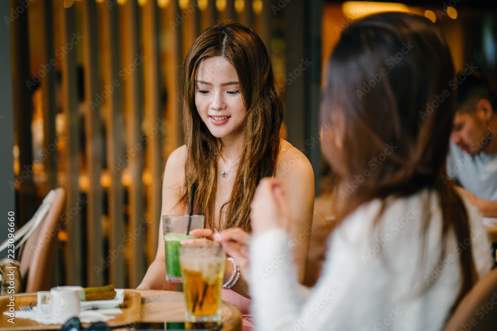 Two young and attractive Asian Chinese women friends are catching up with one another in the day in a cafe. They are both enjoying a cold beverage with one another. The women are fashionably dressed.