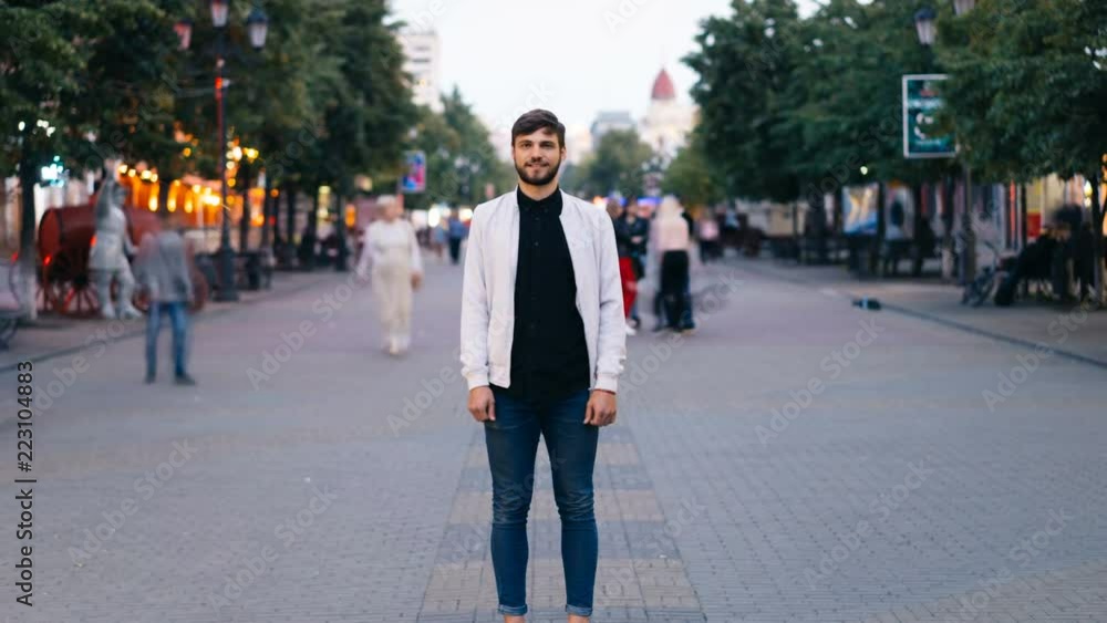 Zoom out time-lapse of confident young man looking at camera standing in city center in pedestrian street while men and women are walking around him in hurry. Zoom out time-lapse of confident young man looking at camera standing in city center in pedestrian street while men and women are walking around him in hurry.