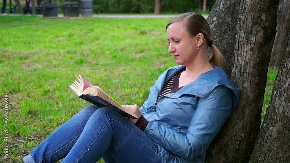 A middle-aged woman sitting on the grass near a tree and reading a book. A middle-aged woman sitting on the grass near a tree and reading a book.