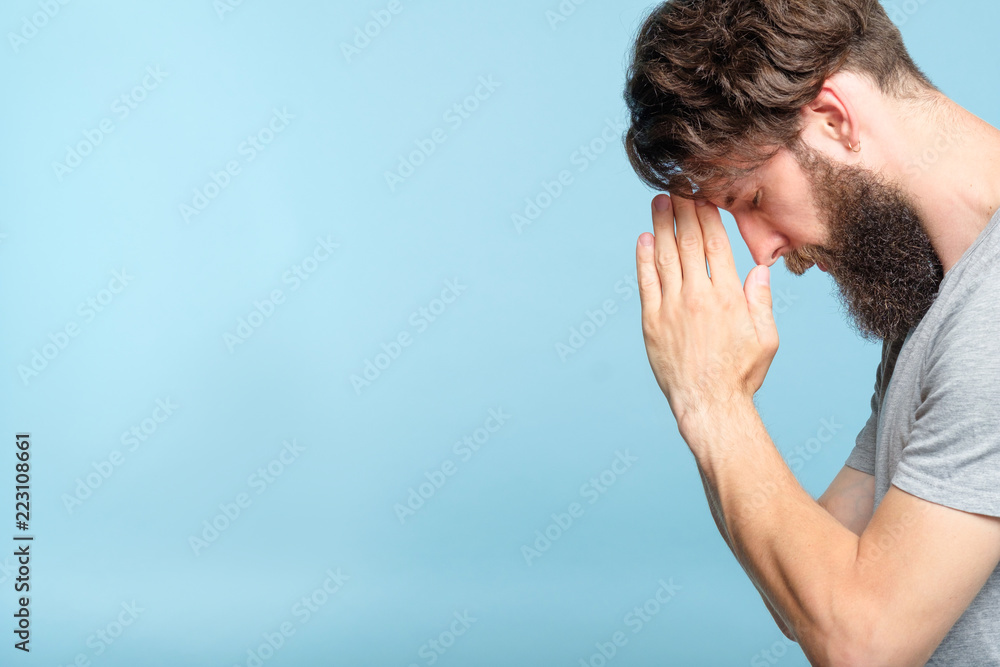 Fototapeta premium man praying. hands touching forehead. spirituality faith and belief concept. portrait of a bearded guy on blue background.