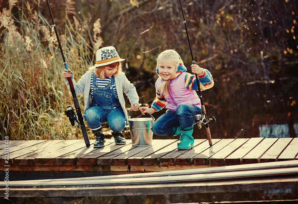 Two little girls with fishing rods, sitting on a wooden pontoon and  bragging of caught fish Stock Photo