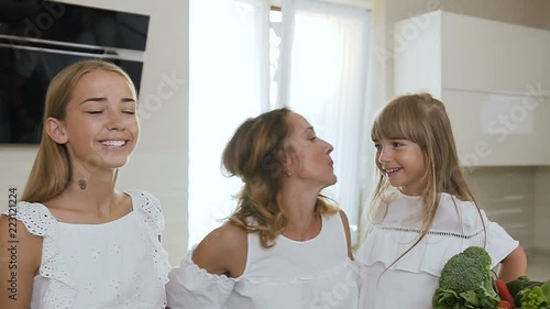 Portrait of a beautiful mother and her cute daughters who kisses in cheeks and hugging each otherd in the kitchen at home. Happy family, healthy, mom with daughters photo