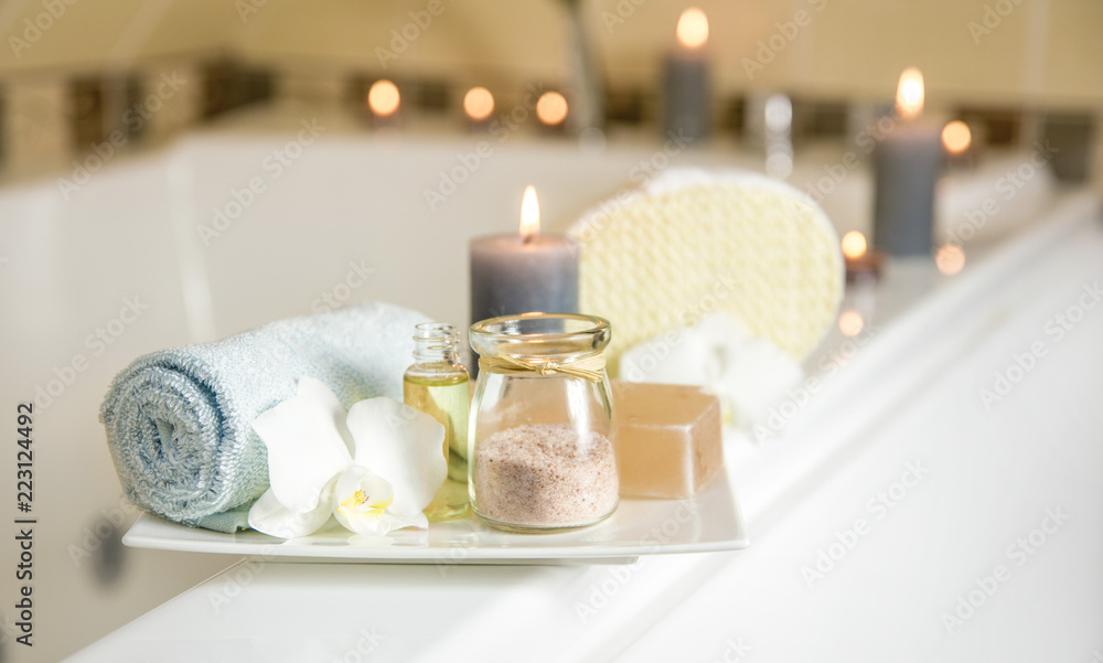 White ceramic tray with home spa supplies in home bathroom for relaxing  rituals. Candlelight, salt soap bar, bath salt in jar, massage, bath oil in  bottle, blue rolled towel, natural sponge. Stock-Foto
