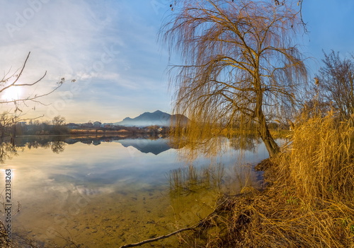Autumn pond with a reflected mountain. Willows on the shore of the pond. The village at the foot of the mountain. Autumn sunset.