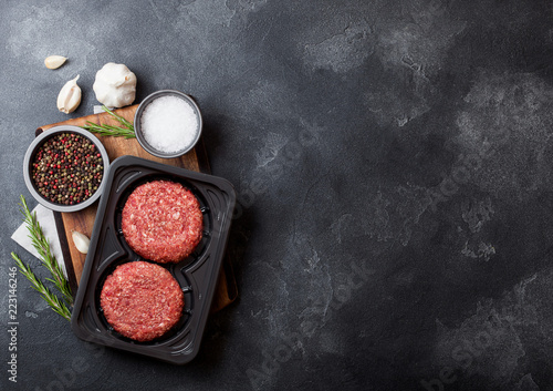 Plastic tray with raw minced homemade beef burgers with spices and herbs. Top view and space for text on stone kitchen table background with tomatoes salt and pepper.