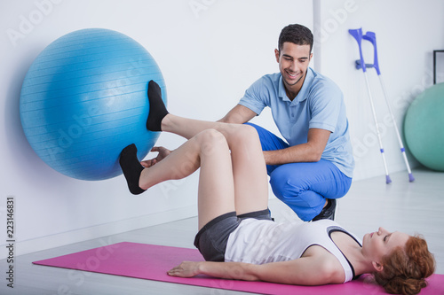 Smiling physiotherapist supporting woman exercising with rehabilitation ball