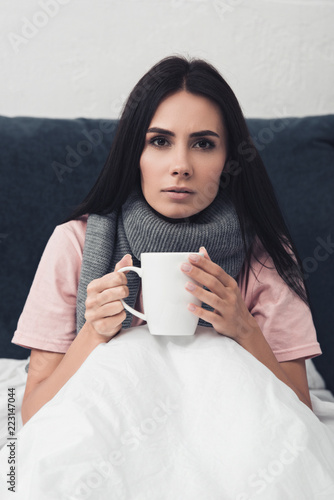 sick young woman in bed holding cup of hot tea while sitting in bed and looking at camera