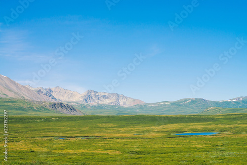 Giant mountains with snow above green valley under clear blue sky. Meadow with rich vegetation and lakes of highlands in sunlight. Amazing sunny mountain landscape of majestic nature.