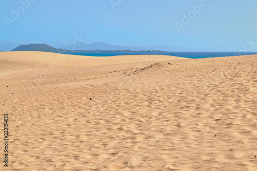 Spectacular paradisiacal landscape of golden sand dunes and blue sea with mountains in background in Corralejo Natural Park, Fuerteventura, Canary Islands, Spain