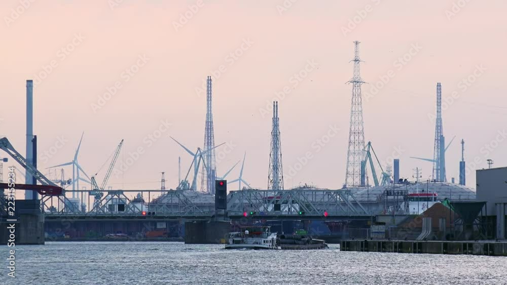 Beautiful view of marine cargo terminal on Scheldt river. Seaport area with cranes, bridge and silhouettes of rotating wind turbines on background. Loaded barge delivers freight. Antwerp, Belgium Beautiful view of marine cargo terminal on Scheldt river. Seaport area with cranes, bridge and silhouettes of rotating wind turbines on background. Loaded barge delivers freight. Antwerp, Belgium
