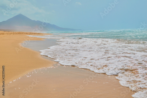 Landscape of turquoise water beach and stone mountain with golden sand and sea foam on the shore at Cofete, Fuerteventura, Canary Islands, Spain