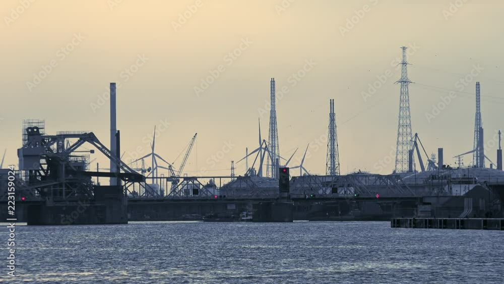 Futuristic scenery with bridge over Scheldt river, waterway traffic signal, dock cranes, buildings of Antwerp marine terminal or sea port and silhouettes of rotating wind turbines. Belgium Futuristic scenery with bridge over Scheldt river, waterway traffic signal, dock cranes, buildings of Antwerp marine terminal or sea port and silhouettes of rotating wind turbines. Belgium