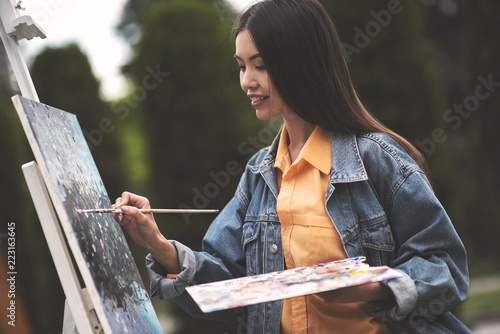 Artist painting on easel in studio outdoor. Girl paints field flowers with brush. Female painter seen from behind. Garden home interior for handmade crafts. Hobby and rest, relaxing weekend