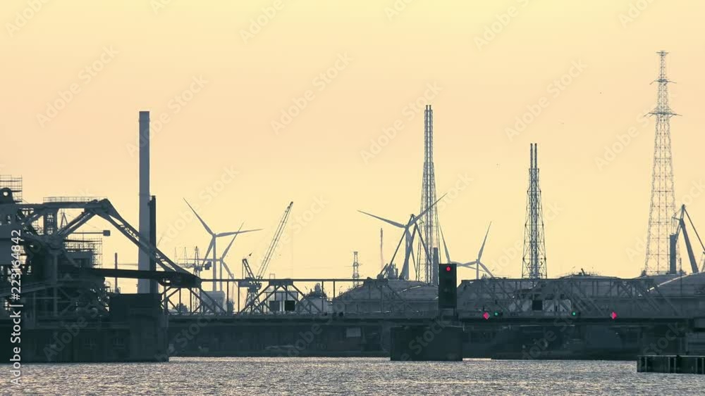 Futuristic industrial scenery with bridge at marine cargo terminal. Silhouettes of cranes and rotating wind turbines against beautiful sunset sky. Seaport area at evening. Antwerp, Belgium