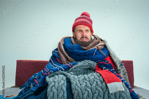Bearded sick man with flue sitting on sofa at home or studio covered with knitted warm clothes. Illness, influenza concept. Relaxation at Home. Healthcare Concepts.