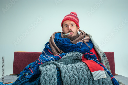 Bearded sick man with flue sitting on sofa at home or studio with thermometer covered with knitted warm clothes. Illness, influenza concept. Relaxation at Home. Healthcare Concepts.