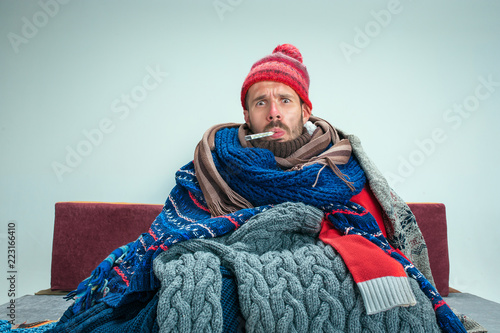Bearded sick man with flue sitting on sofa at home or studio with thermometer covered with knitted warm clothes. Illness, influenza concept. Relaxation at Home. Healthcare Concepts.