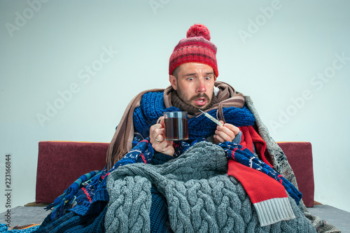 Bearded sick man with flue sitting on sofa at home or studio with cup of tea covered with knitted warm clothes. Illness, influenza concept. Relaxation at Home. Healthcare Concepts.