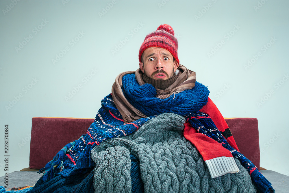 Bearded sick man with flue sitting on sofa at home or studio covered with knitted warm clothes. Illness, influenza, pain concept. Relaxation at Home. Healthcare Concepts. - obrazy, fototapety, plakaty 