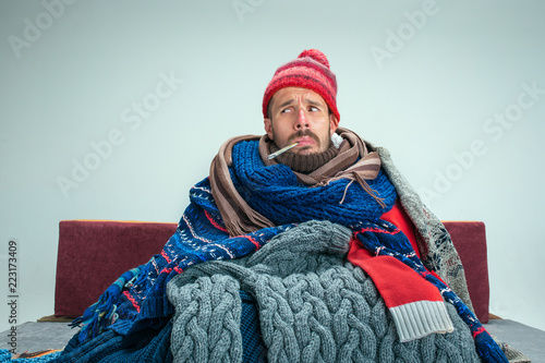 Ταπετσαρία Bearded sick man with flue sitting on sofa at home or studio with thermometer covered with knitted warm clothes