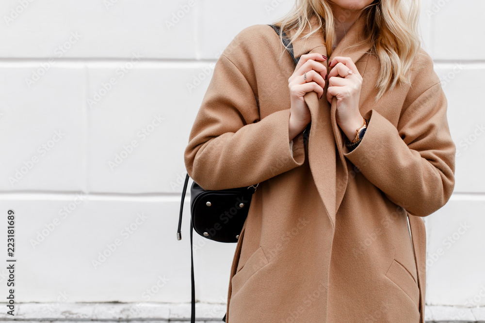 Beautiful young stylish blonde woman wearing beige coat and black backpack posing near white street wall. Trendy casual outfit. Street fashion. Details of everyday look. - obrazy, fototapety, plakaty 