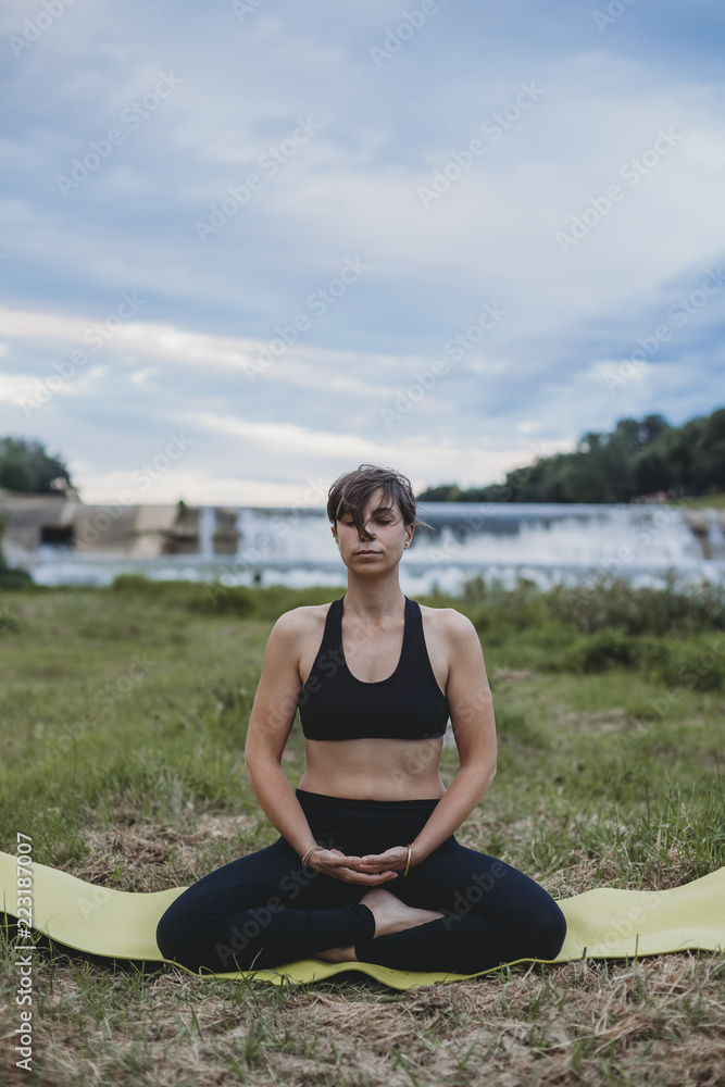 Young beautiful woman doing yoga exercises in park on the bank river in the summer