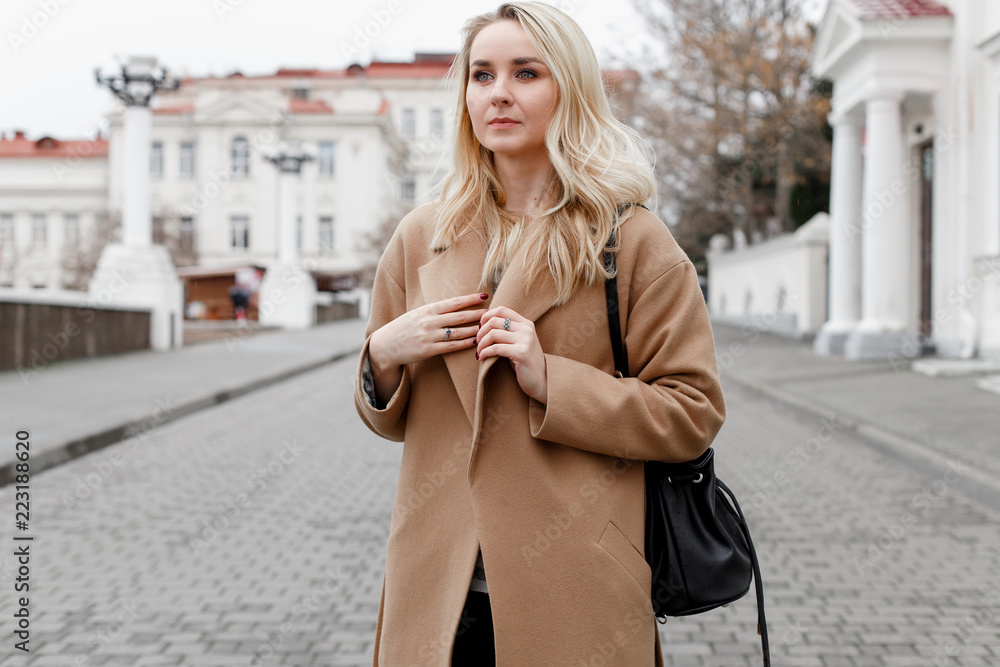 Beautiful young stylish blonde woman wearing beige coat and black backpack  walking through the city streets. Trendy casual outfit. Street fashion.  Stock Photo | Adobe Stock